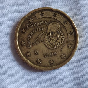 May include: A gold-colored Spanish 1 Euro coin featuring a portrait of Miguel de Cervantes, the author of Don Quixote, with the inscription "ESPAÑA" and "Cervantes" around the portrait. The coin also has the year "1999" and the letter "M" on the bottom.