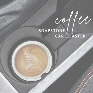 May include: A white soapstone car coaster with the text "coffee" and "SOAPSTONE CAR COASTER" in a car cup holder. A cup of coffee with latte art is sitting on the coaster.
