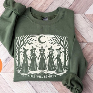 Puede incluir: Sudadera verde con un gráfico blanco de cuatro brujas en un bosque con el texto "Girls will be girls".