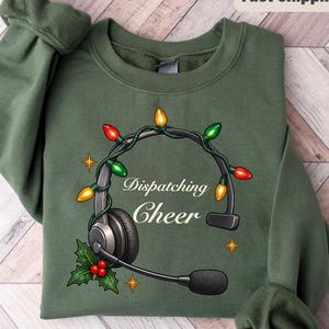 May include: A military green crewneck sweatshirt featuring a graphic of a black headset wrapped in colorful Christmas lights. The text "Dispatching Cheer" is printed in white script in the center, accompanied by holly leaves and berries.