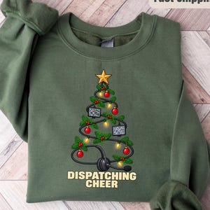 May include: A military green crewneck sweatshirt featuring a graphic of a Christmas tree formed by a black telephone headset cord. The tree is decorated with red ornaments, yellow lights, a gold star, and two computer screens. The text reads DISPATCHING CHEER.