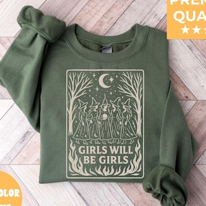 May include: A dark green sweatshirt with a graphic featuring a group of witches under a crescent moon and the text "GIRLS WILL BE GIRLS". The design is in a rectangular frame with trees and flames.