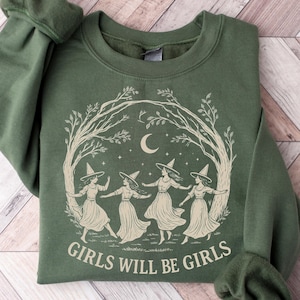 Puede incluir: Sudadera verde con un gráfico de cuatro brujas bailando bajo una luna creciente y estrellas, enmarcado por ramas de árboles. El texto "GIRLS WILL BE GIRLS" está impreso debajo. La sudadera tiene mangas largas.