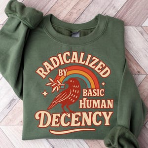 May include: Green sweatshirt with the text "RADICALIZED BY BASIC HUMAN DECENCY" in a retro font. A red bird with a sprig and a rainbow graphic are also featured. The sweatshirt is folded on a wooden surface.