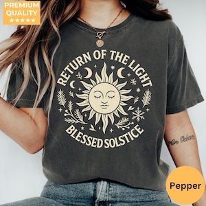 以下が含まれることがあります： クリーム色の太陽のグラフィック、三日月、そして「RETURN OF THE LIGHT BLESSED SOLSTICE」の文字が入った濃い灰色のTシャツ。 ゆったりとしたフィット感と半袖です。 右下には「Pepper」という文字が入った黄色の円があります。