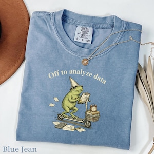 Puede incluir: Camiseta azul vaquero con la frase "Off to analyze data" y un gráfico de una rana en un scooter, con un sombrero de mago y una carpeta. La camiseta tiene una etiqueta "Comfort Colors". Un collar de oro está drapeado sobre la camiseta.
