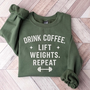 May include: An olive green sweatshirt with the text "DRINK COFFEE. LIFT WEIGHTS. REPEAT" in white. The design includes two small star shapes and a barbell icon. The sweatshirt is folded on a light brown wooden surface.