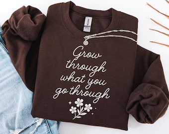 May include: A dark chocolate-colored sweatshirt with the phrase "Grow through what you go through" in white script. The design includes a floral graphic. A silver necklace is draped across the front. The words "Dark Chocolate" are at the bottom.