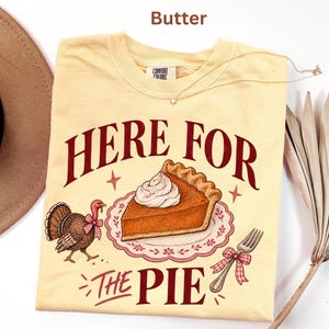 May include: A butter-coloured t-shirt featuring a graphic of a slice of pumpkin pie on a plate, a turkey with a red and white chequered bow, and a fork with a matching bow. The text reads HERE FOR THE PIE in dark red, with small star accents.
