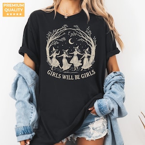 May include: A black short-sleeved t-shirt featuring a cream-coloured graphic of four figures wearing pointed hats dancing in a circle under a crescent moon and tree branches, with the text GIRLS WILL BE GIRLS printed in a bold serif font below.