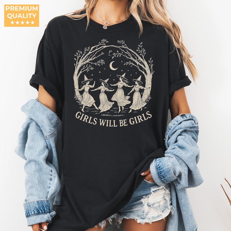 May include: A black short-sleeved t-shirt featuring a cream-coloured graphic of four figures wearing pointed hats dancing in a circle under a crescent moon and tree branches, with the text GIRLS WILL BE GIRLS printed in a bold serif font below.
