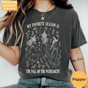May include: A charcoal grey t-shirt featuring a white graphic of three figures wearing witch hats and long dresses surrounded by floral elements. The text reads MY FAVORITE SEASON IS THE FALL OF THE PATRIARCHY in a white serif font.
