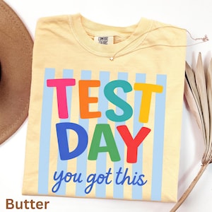 Könnte beinhalten: Ein hellgelbes T-Shirt mit den Worten "TEST DAY you got this" in bunten Buchstaben. Das Shirt hat hellblaue vertikale Streifen hinter dem Text. Die Marke "Comfort Colors" ist auf dem Etikett sichtbar.