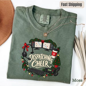 May include: A moss green t-shirt featuring a graphic of a holiday wreath made of computer monitors, a headset, a keyboard, a radio, a coffee cup, and a Christmas tree. The text "DISPATCHING CHEER" is printed in the center in a cream font.
