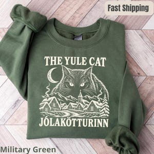 Puede incluir: Una sudadera de cuello redondo color verde militar con un estampado gráfico blanco de una cara de gato grande sobre un paisaje de pueblo nevado. El texto THE YULE CAT y JÓLAKÖTTURINN está impreso en blanco sobre y bajo la ilustración.