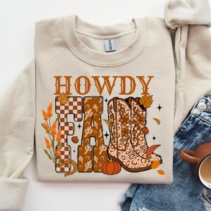 May include: Beige crew neck sweatshirt with a fall-themed graphic. The design features the word "HOWDY" in brown letters above the word "FALL". The letters are decorated with a checkered pattern, cacti, and cowboy boots. A pumpkin and autumn leaves complete the design.