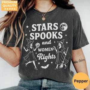 Può includere: T-shirt grigio scuro con la scritta bianca "STARS, SPOOKS, and WOMEN'S Rights". Il design include illustrazioni di streghe, stelle, una luna e un pipistrello. La maglietta ha un aspetto vintage.