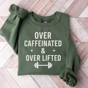 May include: An olive green sweatshirt with the text "OVER CAFFEINATED & OVER LIFTED" in white. The design includes two star symbols and a barbell graphic. The sweatshirt is folded on a light brown wooden surface.