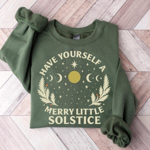 Könnte beinhalten: Ein olivgrünes Sweatshirt mit dem weißen Text "HAVE YOURSELF A MERRY LITTLE SOLSTICE". Das Design umfasst eine gelbe Sonne, Halbmonde, Sterne und botanische Elemente.
