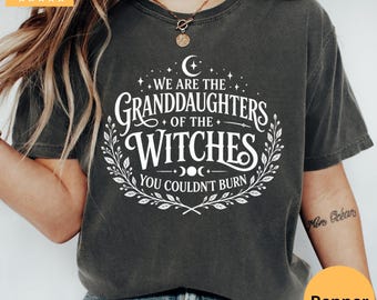 May include: A charcoal gray short-sleeve t-shirt featuring white text that reads WE ARE THE GRANDDAUGHTERS OF THE WITCHES YOU COULDN'T BURN, surrounded by a crescent moon, stars, and a leafy vine border design printed on the front of the garment.