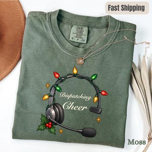 May include: A moss green short-sleeve t-shirt featuring a graphic of black headphones wrapped in colorful Christmas lights. The text reads Dispatching Cheer, accompanied by holly leaves and berries. The shirt has a Comfort Colors brand label.