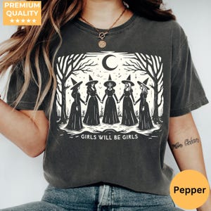 Op de afbeelding: Een antracietgrijs t-shirt met korte mouwen en een crèmekleurige afbeelding van vijf figuren in puntmutsen en lange jurken die elkaars hand vasthouden onder een maansikkel en bomen, met de tekst GIRLS WILL BE GIRLS eronder gedrukt.