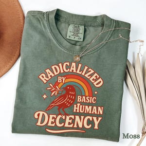 May include: A moss green t-shirt featuring the slogan "RADICALIZED BY BASIC HUMAN DECENCY" in a retro style. The design includes a bird with a branch, a rainbow, and stars. A silver necklace with a pendant is placed on the shirt.