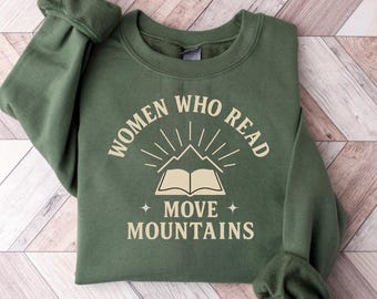 May include: An olive green sweatshirt with the text "WOMEN WHO READ MOVE MOUNTAINS" in a cream-colored font. The design includes an open book, mountain graphic, and star accents. The sweatshirt is folded on a wooden surface.