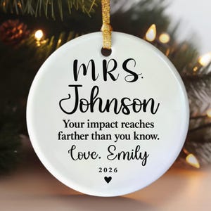 May include: A round white ceramic ornament with a gold ribbon hanger. The text reads: MRS. Johnson, Your impact reaches farther than you know. Love, Emily, 2026, with a small black heart icon centered at the bottom of the ornament.