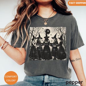 May include: A dark gray t-shirt with a graphic print of four witches in black dresses and pointed hats. The witches are standing in a circle and the text "Girls Will Be Girls." is printed below them.