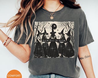 May include: A dark grey t-shirt featuring a graphic print of four witches in black dresses and pointed hats. The witches are standing in a circle and the text "Girls Will Be Girls." is printed below them.