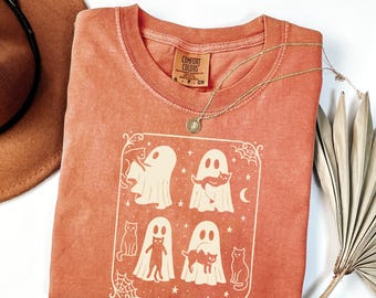 May include: A coral-colored Comfort Colors t-shirt featuring a cream-colored graphic of ghosts and cats within a decorative frame. The shirt has a crew neck and short sleeves. A gold necklace with a pendant is draped across the shirt.