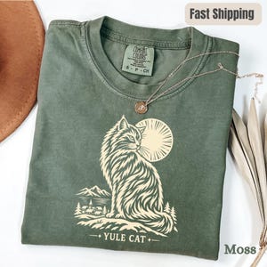 Puede incluir: Una camiseta de manga corta de color verde oliva con un gráfico de color crema de un gato de pelo largo sentado frente a un sol, con un paisaje de montaña y pueblo debajo. El texto YULE CAT está impreso en la parte inferior.