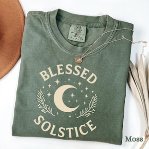 以下が含まれることがあります： モスグリーンのTシャツ。クリーム色の「BLESSED SOLSTICE」の文字と三日月と星のデザイン。 金色のネックレスが折りたたまれたシャツの上にかけられています。 シャツにはタグが付いており、サイズはSです。