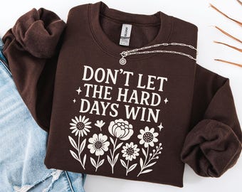 May include: Dark chocolate-colored sweatshirt with the text "DON'T LET THE HARD DAYS WIN" and a floral design in white. A silver necklace with a pendant is draped across the sweatshirt. The bottom of the image shows the words "Dark Chocolate".