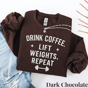 May include: A dark chocolate-colored sweatshirt with the text "DRINK COFFEE. LIFT WEIGHTS. REPEAT" in white. The sweatshirt has a crew neck and long sleeves. A silver necklace with a pendant is draped on the sweatshirt. The words "Dark Chocolate" are at the bottom.