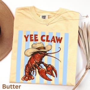 Puede incluir: Camiseta de color mantequilla con un diseño de un cangrejo de río vaquero y el texto "Yee Claw". El diseño presenta un cangrejo de río rojo con un sombrero de vaquero sobre un fondo de rayas azules. Marca Comfort Colors.