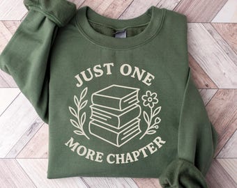 Puede incluir: Sudadera verde oliva con el texto blanco "JUST ONE MORE CHAPTER". El texto está centrado sobre una pila de libros, una flor y hojas. La sudadera tiene mangas largas y cuello redondo.
