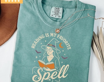 May include: Light green t-shirt with the text "READING IS MY FAVORITE SPELL" and an illustration of a witch reading a book. The shirt has a crew neck and short sleeves. A silver necklace is draped across the shirt.