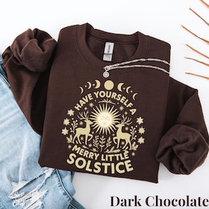 Może przedstawiać: Ciemnobrązowa bluza z kremową grafiką. Grafika przedstawia słońce, jelenie, księżyce, gwiazdy i napis "Have Yourself a Merry Little Solstice". Srebrny naszyjnik z zawieszką w kształcie słońca jest na bluzie.