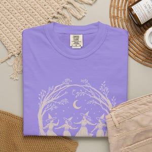 May include: A light purple t-shirt featuring a cream-coloured graphic print of four figures wearing pointed hats dancing under an arch of tree branches with a crescent moon and stars in the background. The inner neck label reads Comfort Colors.