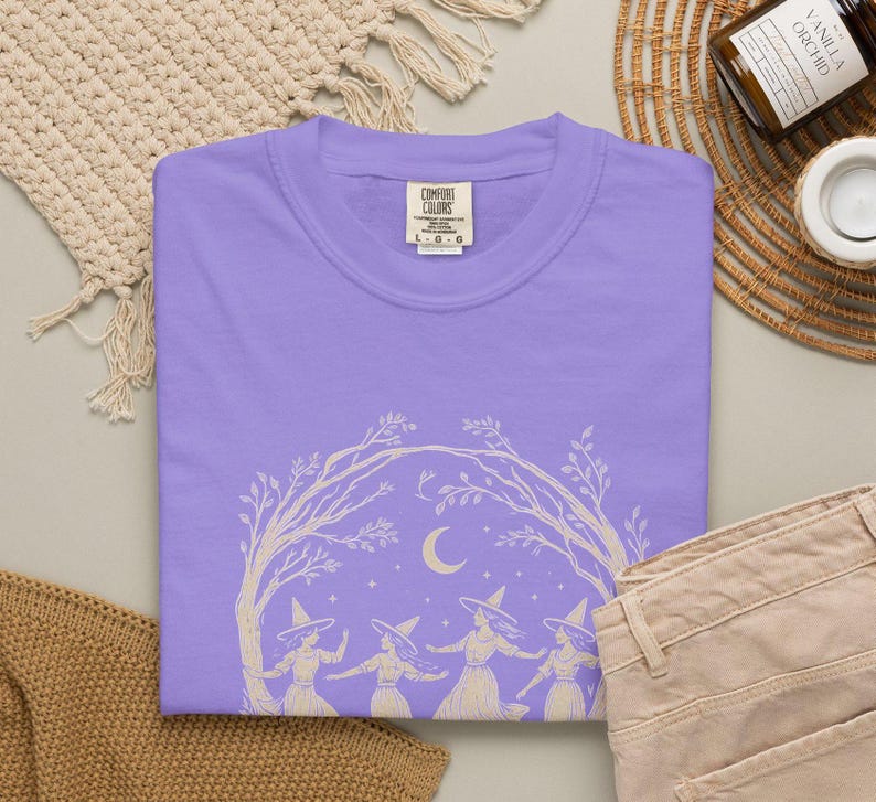 May include: A light purple t-shirt featuring a cream-coloured graphic print of four figures wearing pointed hats dancing under an arch of tree branches with a crescent moon and stars in the background. The inner neck label reads Comfort Colors.