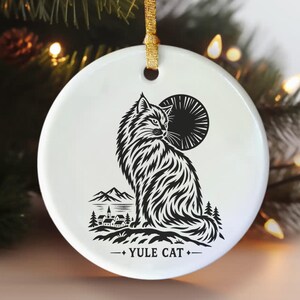 Może przedstawiać: Okrągła biała ceramiczna ozdoba z czarną grafiką przedstawiającą długowłosego kota siedzącego przed słońcem. Pod kotem znajduje się mała wioska z górami oraz napis YULE CAT, zawieszona na złotej wstążce.