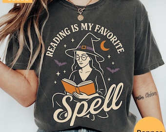 May include: Dark gray t-shirt with the text "Reading is my favorite spell" featuring a witch reading a book. The design includes a witch hat, bats, stars, and a crescent moon. The shirt has a vintage, worn look.