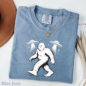 May include: Blue jean colored t-shirt featuring a Bigfoot graphic walking between two UFOs. The Bigfoot is white with black outlines, and the UFOs are white with light beams. The shirt has a Comfort Colors tag and a gold necklace.
