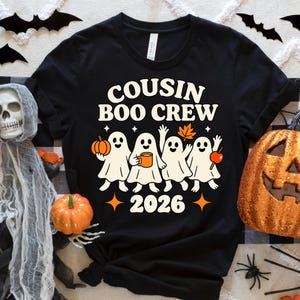 May include: A black short-sleeved t-shirt featuring a graphic of four white ghosts with the text "COUSIN BOO CREW" and "2026" in a cream font. The ghosts are holding an orange pumpkin, a mug, a maple leaf, and a red candy apple.