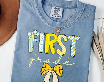 May include: A light blue t-shirt with the words "First Grade" in yellow letters with colorful patterns. The letters are decorated with a yellow bow.