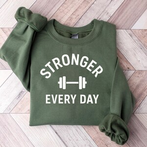 Puede incluir: Una sudadera verde oliva con las palabras "STRONGER EVERY DAY" en letras blancas. Un gráfico de barra con pesas blanco está centrado entre las palabras. La sudadera está doblada sobre una superficie de madera marrón claro.