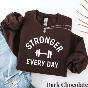 Może przedstawiać: Ciemnobrązowa bluza z napisem "STRONGER EVERY DAY" w białych literach i grafiką sztangi. Srebrny naszyjnik z wisiorkiem jest zarzucony na bluzę. Bluza jest połączona z niebieskimi dżinsami.