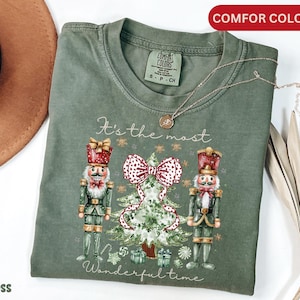 Puede incluir: Una camiseta verde con un diseño navideño con dos figuras de cascanueces que flanquean un árbol de Navidad decorado. El texto "It's the most Wonderful time" está impreso debajo del diseño.